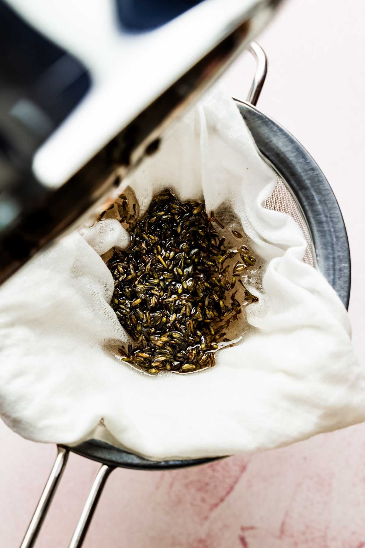 Cooked lavender buds being strained through cheesecloth in a fine mesh strainer.