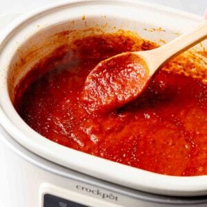 Overhead view of rich red spaghetti sauce in a white slow cooker with a wooden spoon.