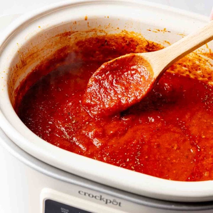 Overhead view of rich red spaghetti sauce in a white slow cooker with a wooden spoon.