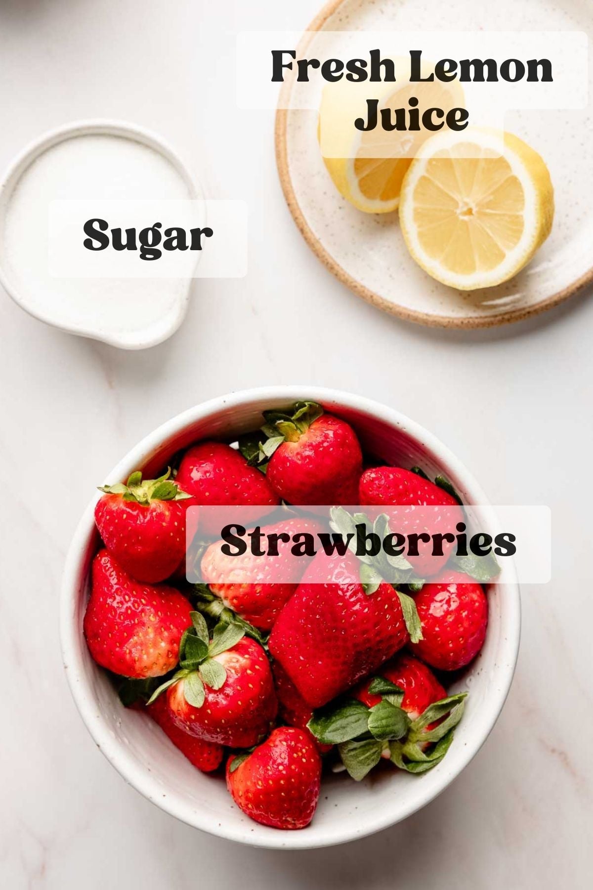 Overhead flat lay of strawberry sauce ingredients in bowls, labeled with fresh strawberries, sugar, and fresh lemon juice, arranged on a white marble surface.