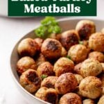 A large shallow white bowl filled with golden-brown baked turkey meatballs, garnished with a sprig of curly parsley. A blurred bowl of pasta and a jar of marinara sauce are visible in the background.