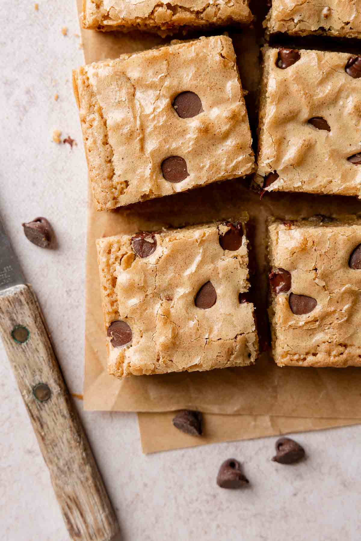 Squares of blondies on parchment paper, showing chocolate chips peeking out the top and shiny surface.