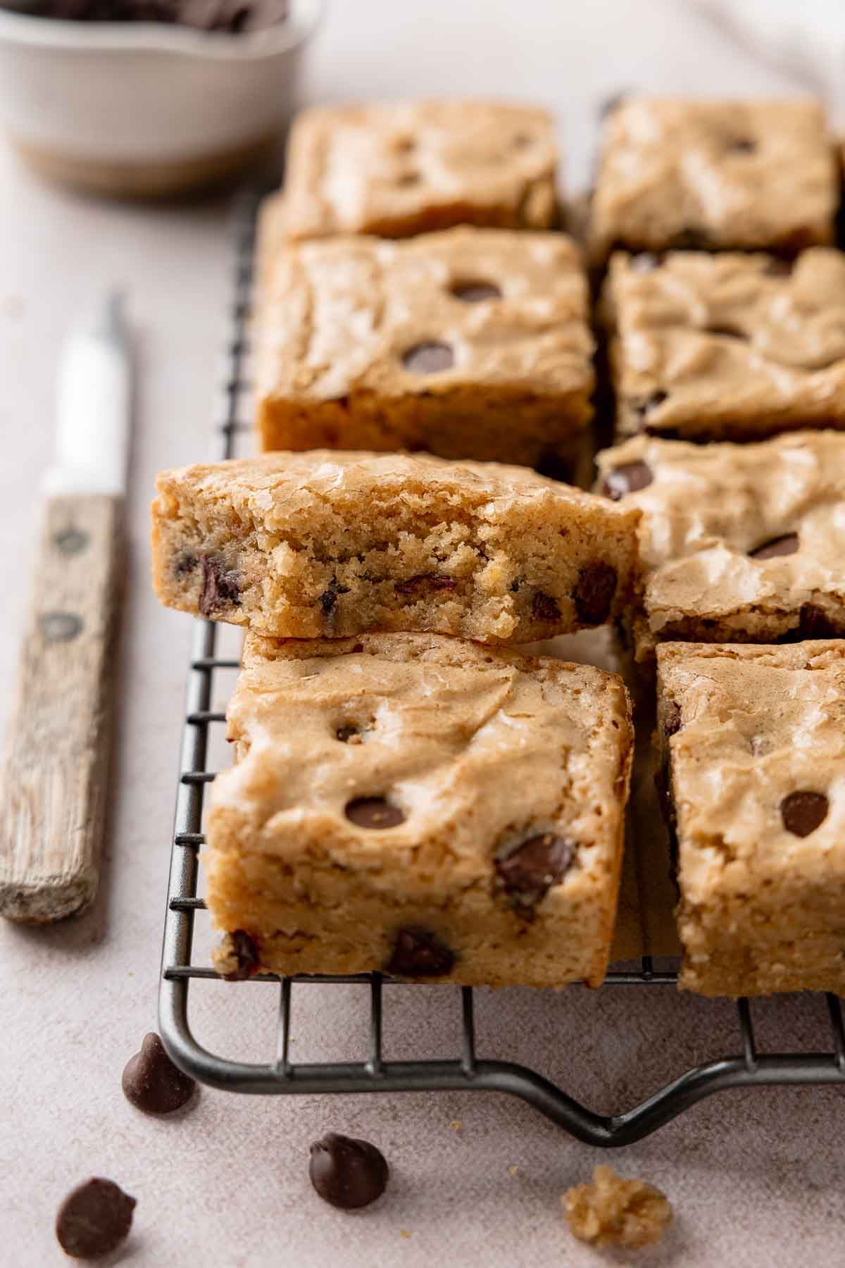 A blondie square with bite taken rests upon another blondie on wire rack lined with more blondies.