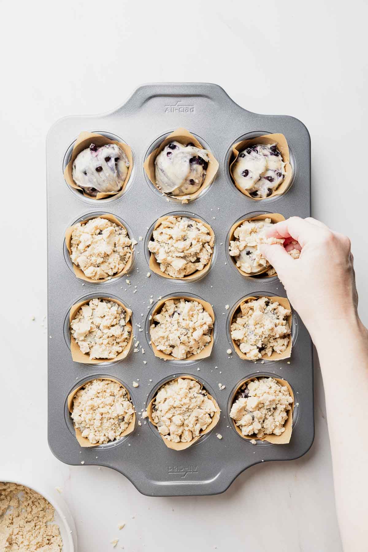 Muffin batter divided amongst parchment paper lined muffin tins, hand topping with streusel.