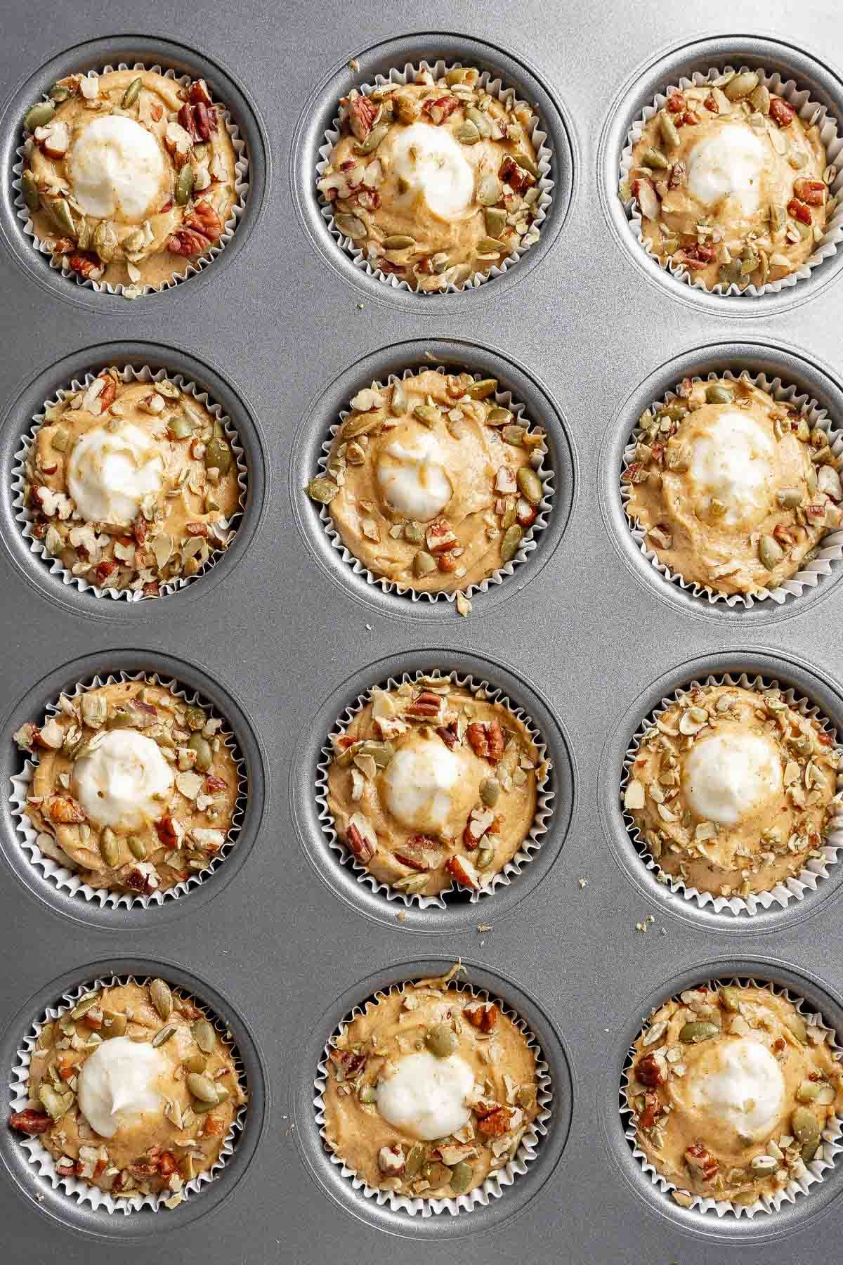 Muffin batter in muffin cups, topped with pecans and pepitas, with a dollop of cream cheese filling in the center of each one.