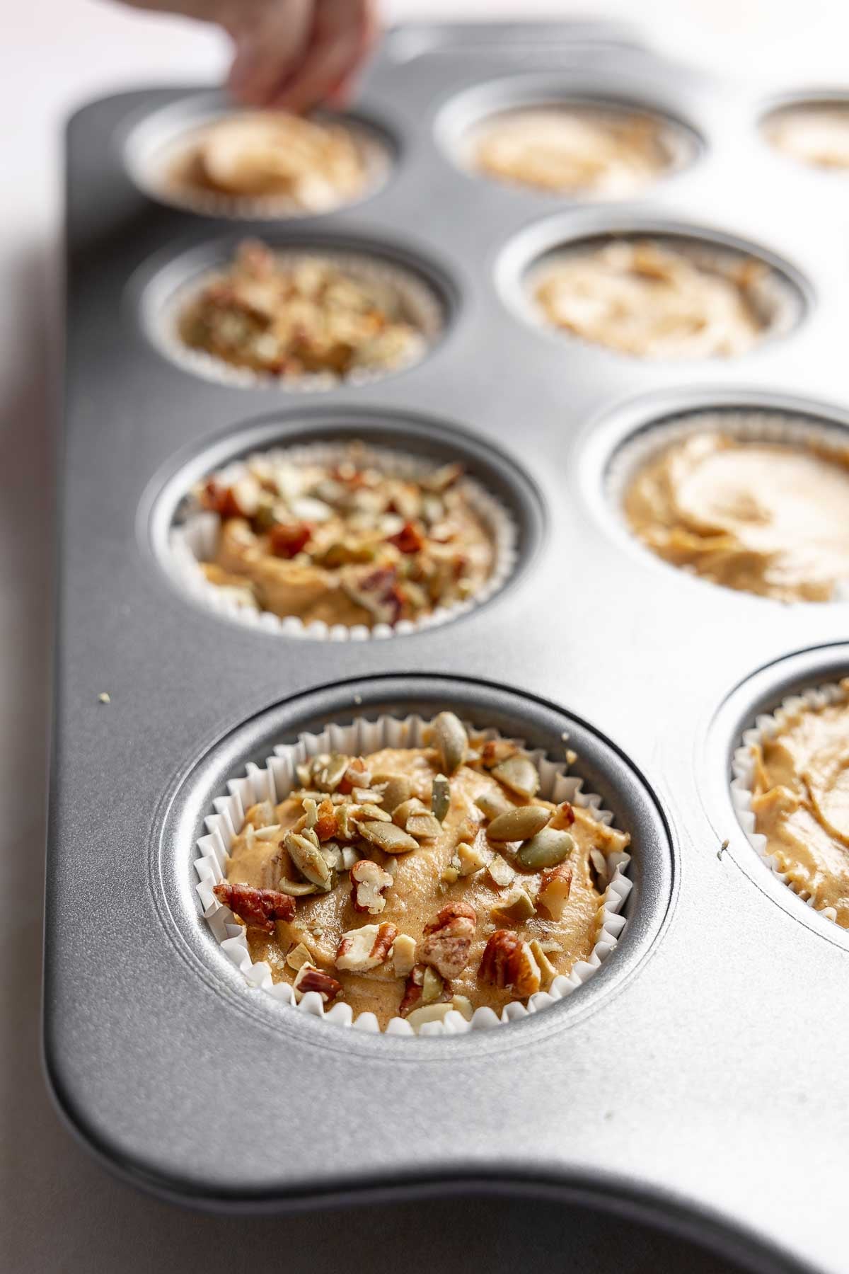 Pumpkin muffin batter in paper lined muffin cups, topped with chopped pepitas and pecans.