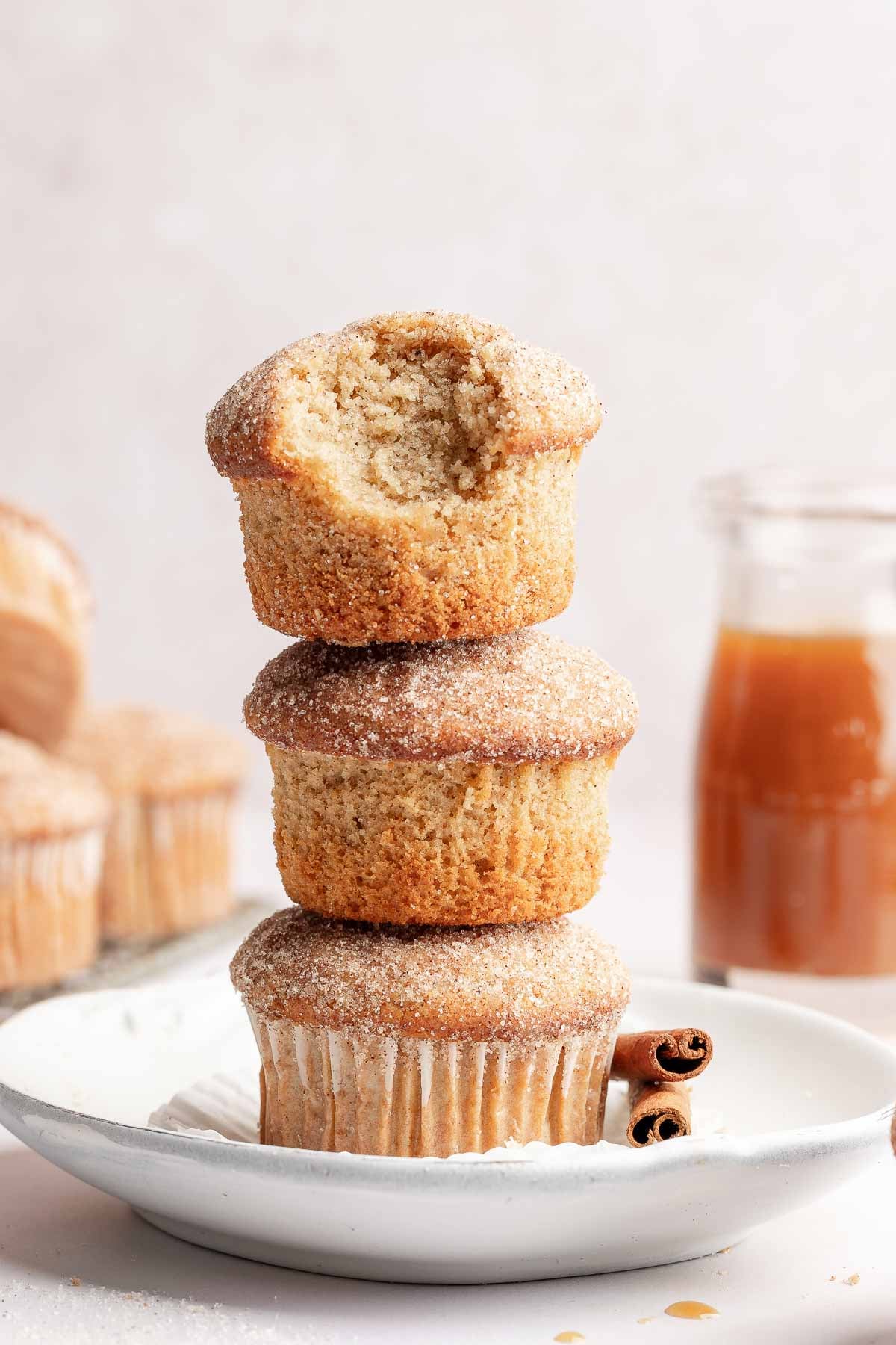 A stack of gluten-free apple cider donut muffins on a small white plate, a jar of apple cider in the background.