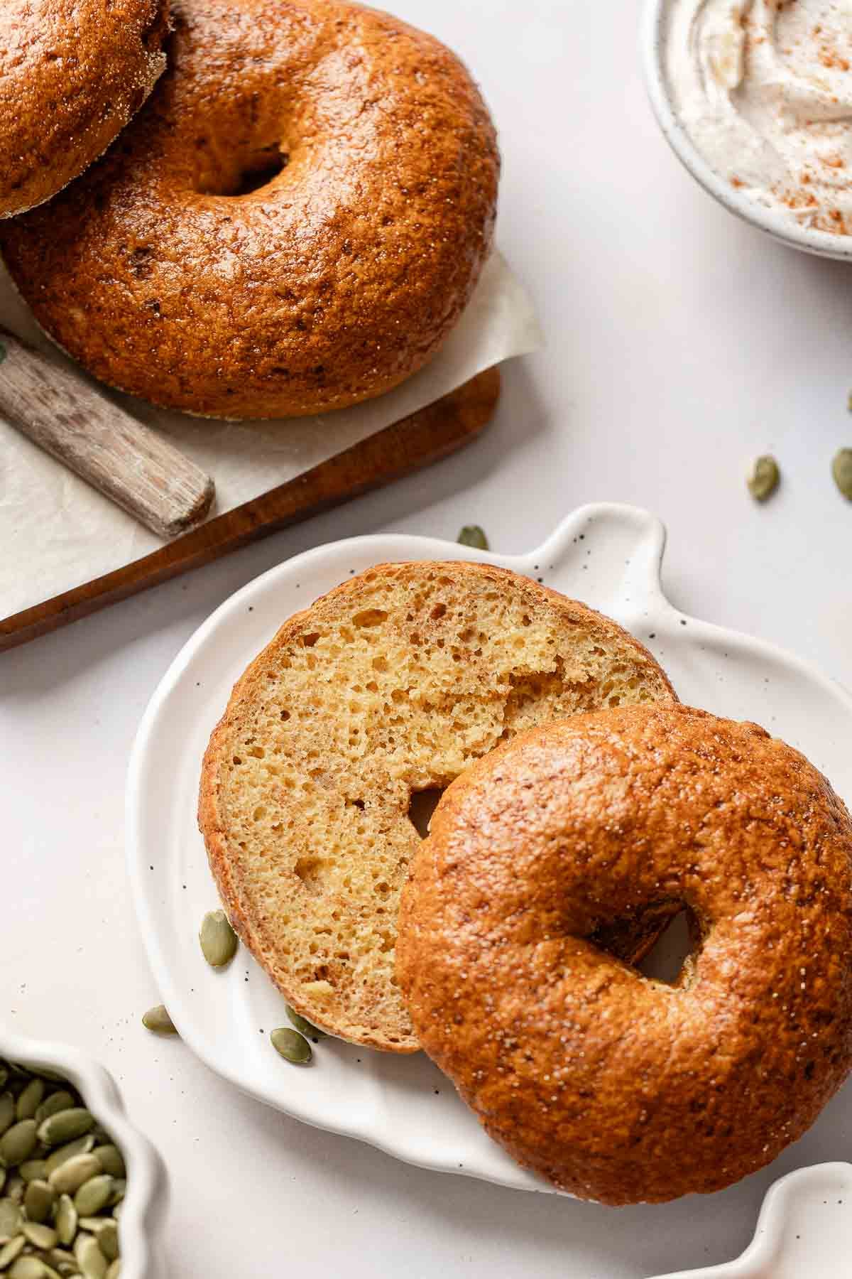 A sliced gluten-free pumpkin bagel showing its airy interior and cinnamon streaks on a white pumpkin-shaped plate.