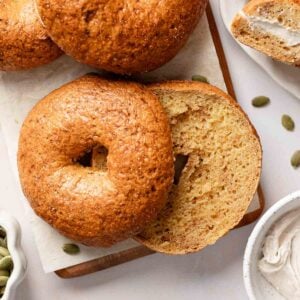 Golden-brown gluten-free pumpkin bagels on a wooden board, with one sliced to show cinnamon streaks and another spread with cream cheese.