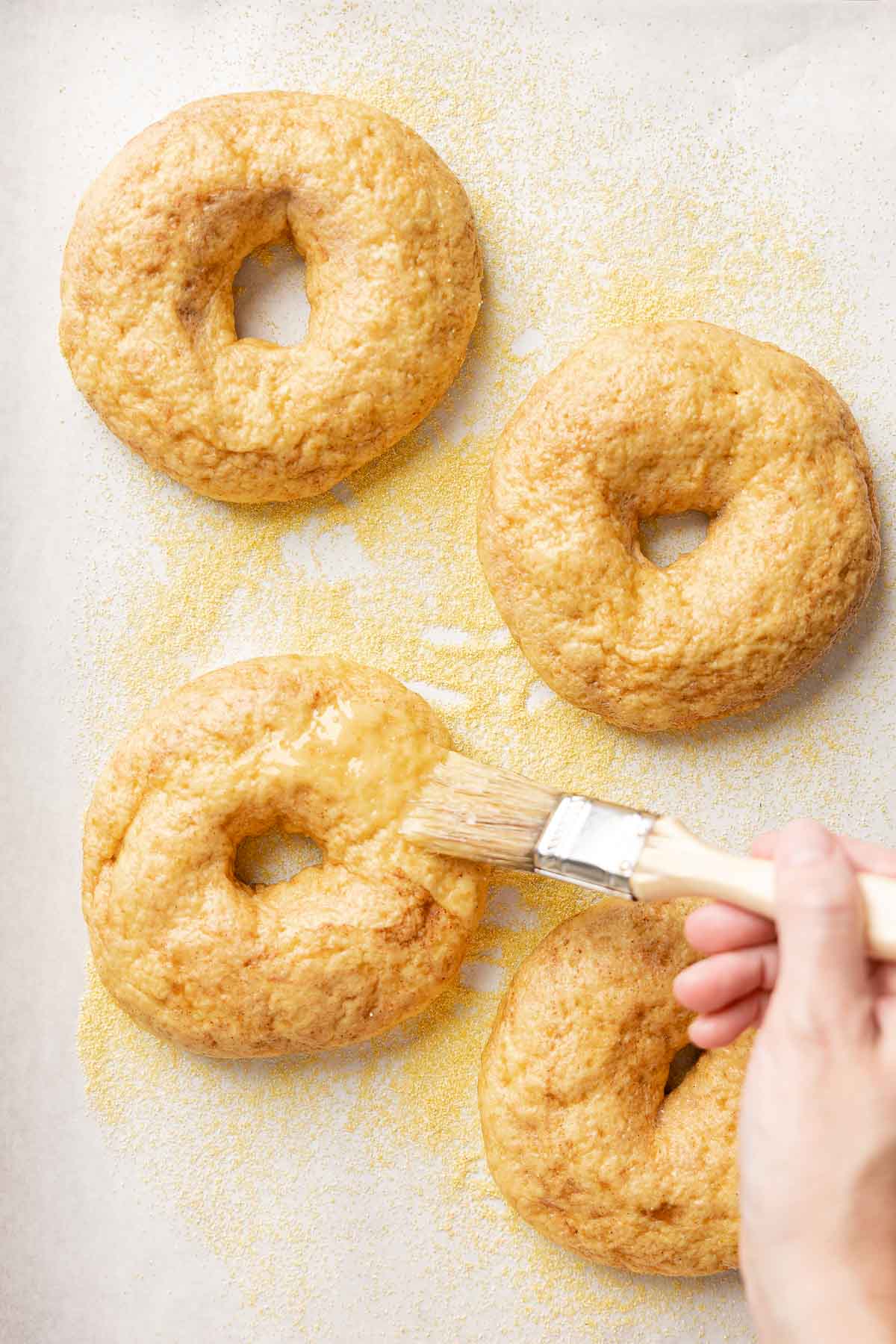 Brushing boiled gluten-free pumpkin bagels with egg wash on cornmeal-dusted parchment before baking.